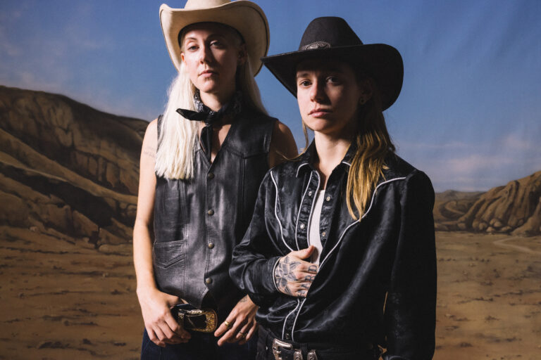 Julien Baker & TORRES tragen Cowboyhüte und stehen vor einer gemalten Landschaft